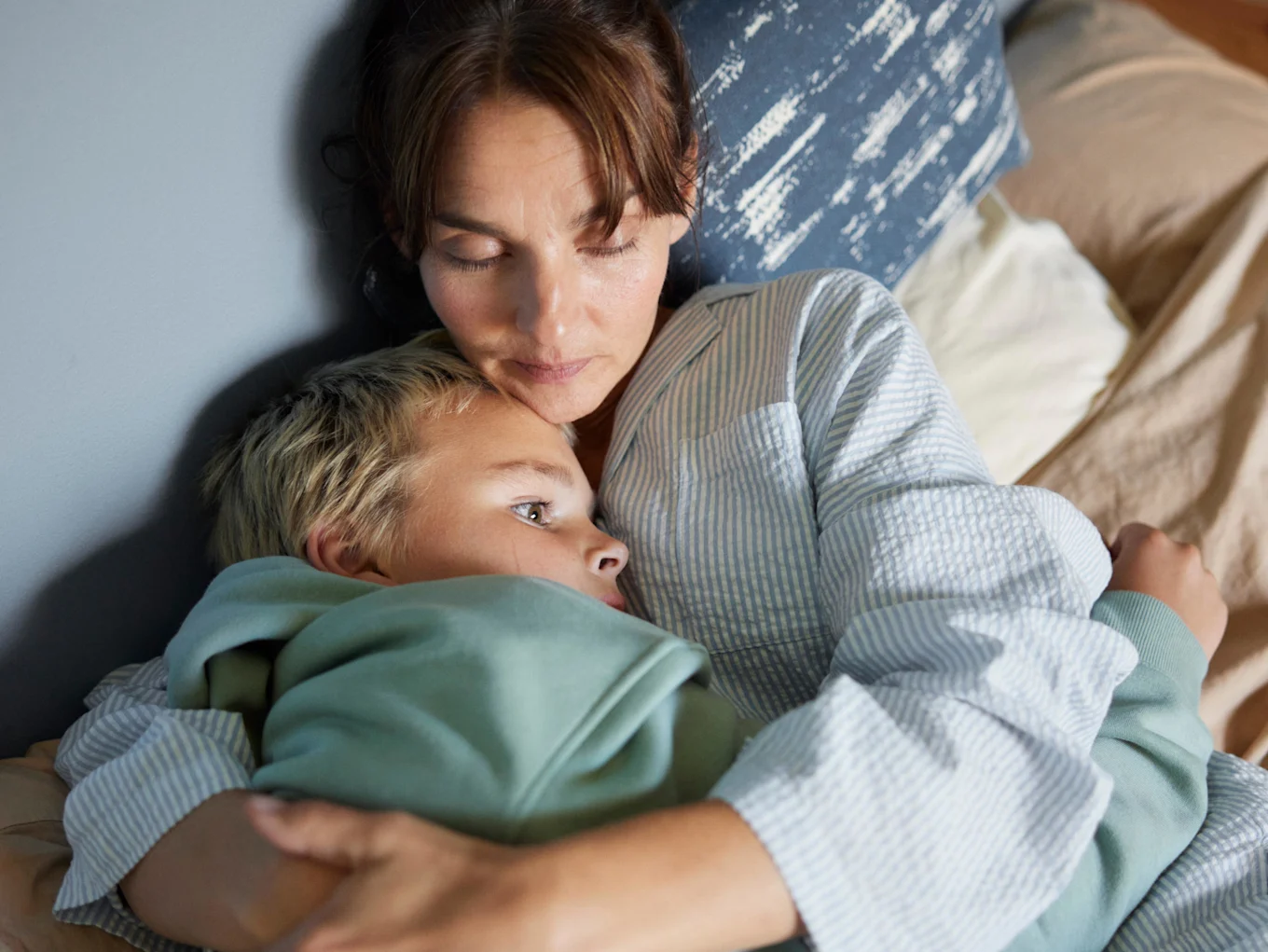 Mamma och son ligger i sängen och vilar och kramas. 