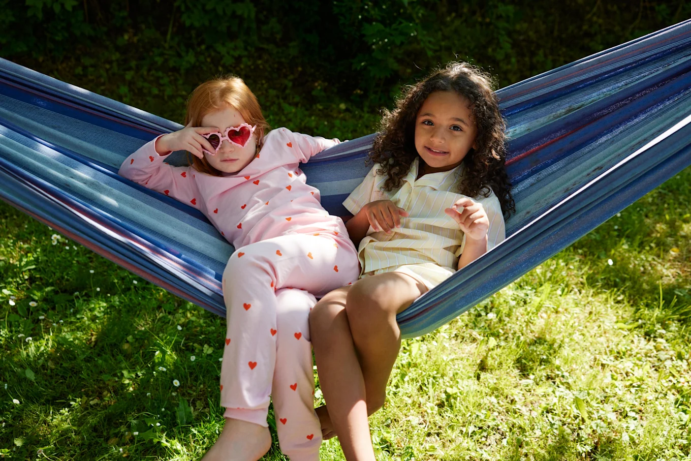 Två flickor i 5-6-årsåldern sitter tillsammans i en hängmatta och tittar mot kameran. Det är sommar och sol