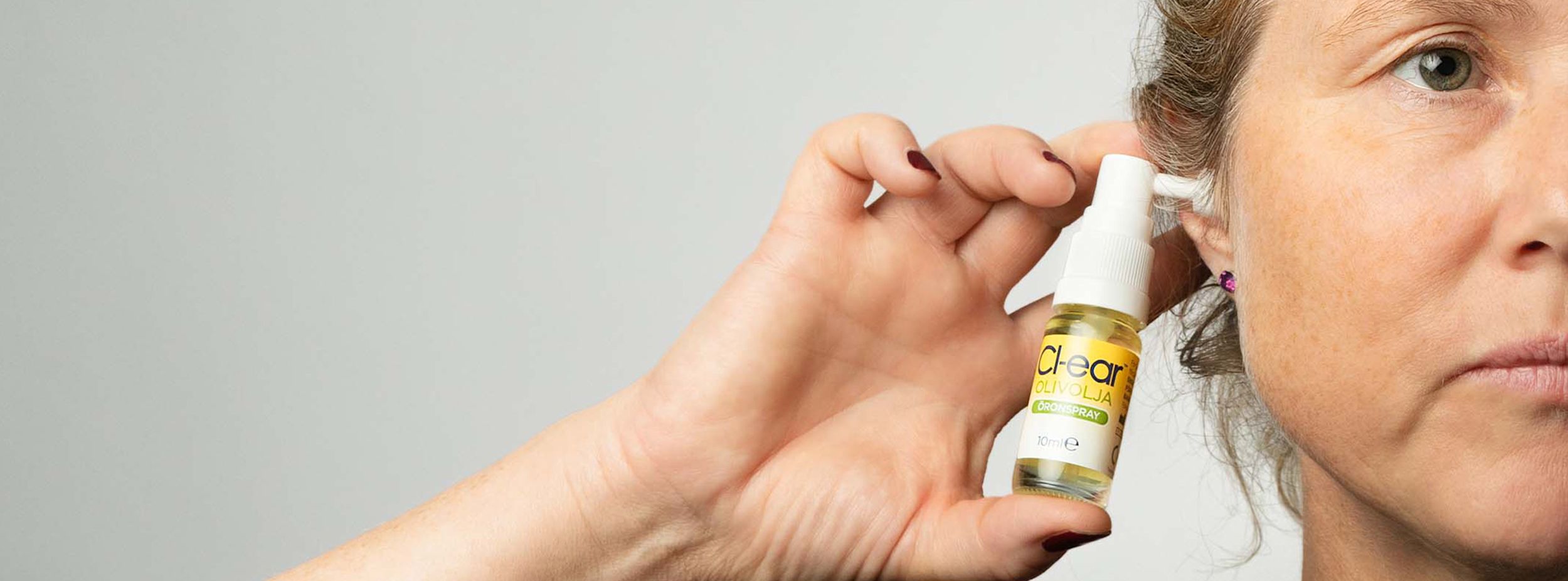 Kvinna håller en flaska Cl-ear olivolja öronspray nära sitt öra mot en ljus bakgrund.