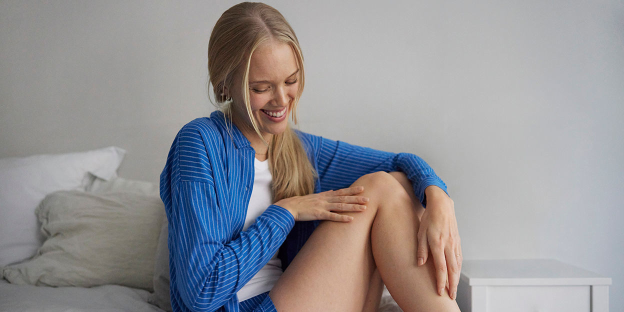 Ung kvinna sitter på sin säng med lång blå och vitrandig skjorta med matchande shorts. Man kan se hennes ben och hon verkar precis ha smörjt in dem.