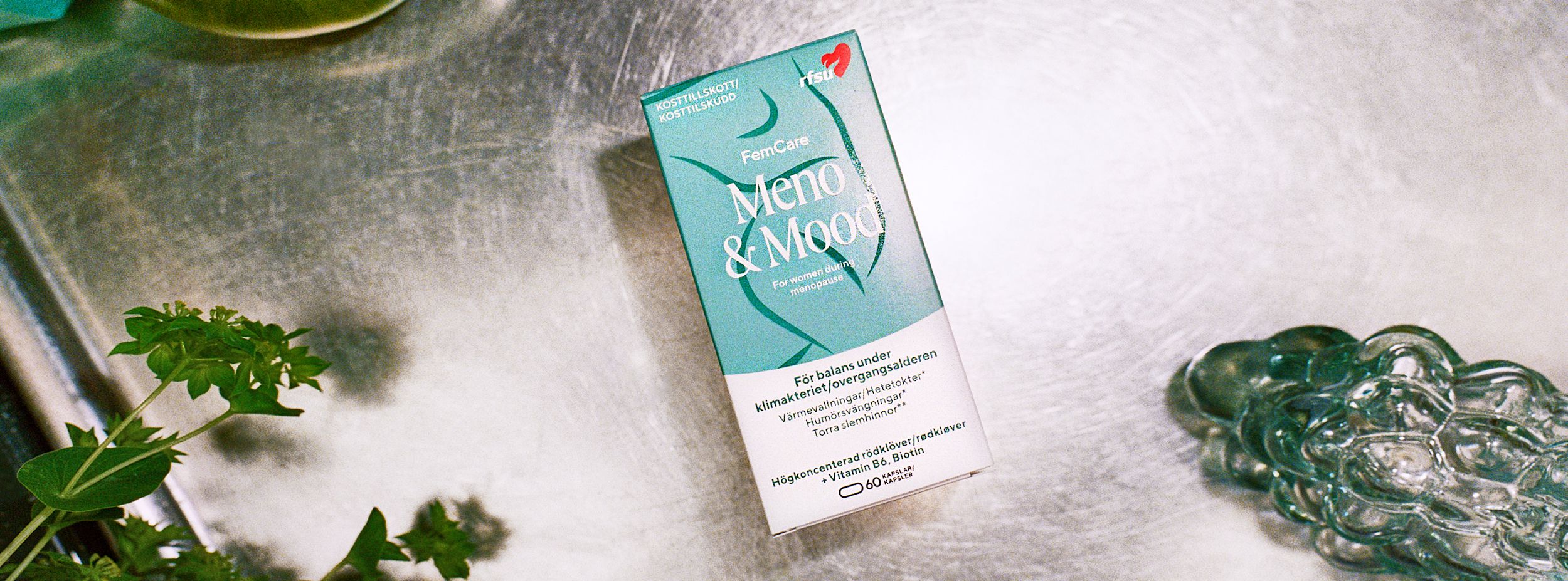 Förpackning av FemCare Meno & Mood kosttillskott ligger på en metallbänk, omgiven av gröna växter och ett glasföremål.