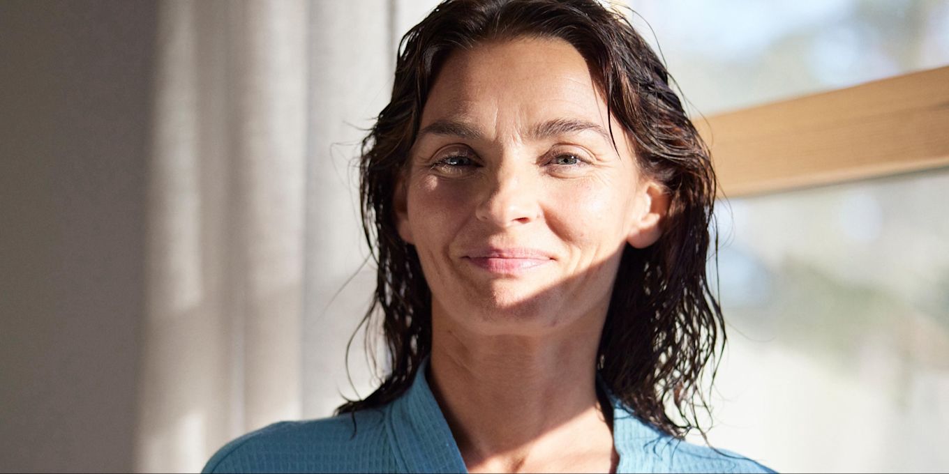 Kvinna står med blött hår i badrummet, iklädd morgonrock.