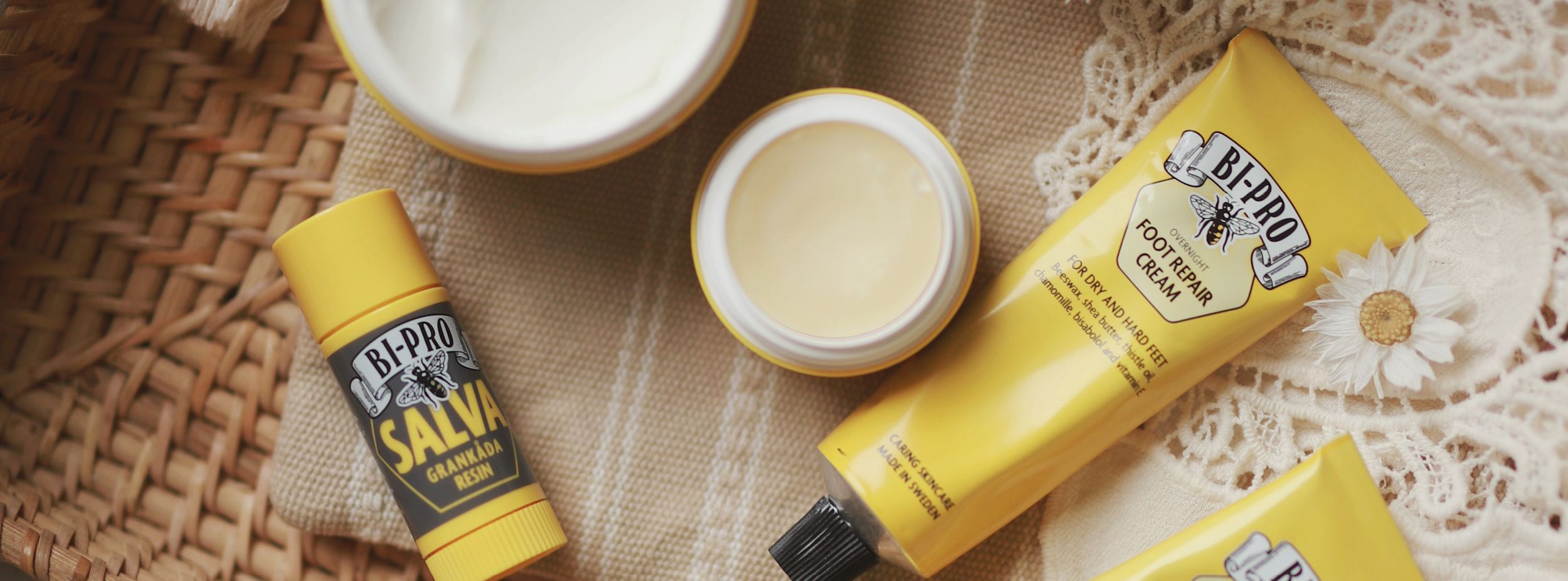 Flera BI-PRO hudvårdsprodukter, inklusive salva och fotkräm, ligger på en beige duk bredvid en vit blomma och korg.