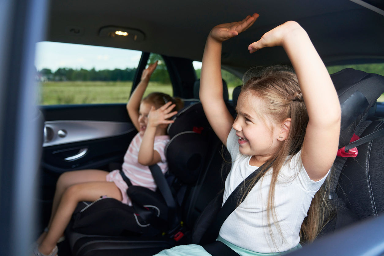 Två barn sitter i baksätet på bilen med armarna uppsträckta och skrattar.