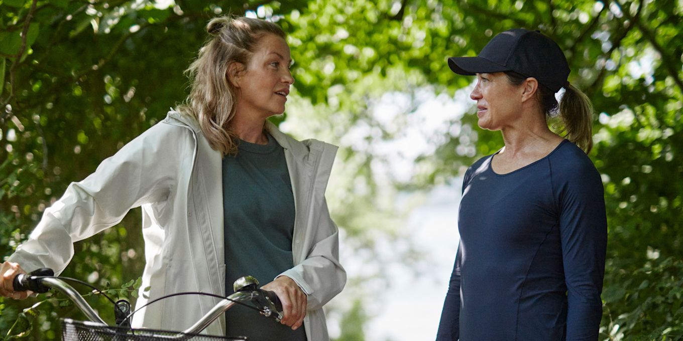 Två kvinnor på promenad står i skogen och pratar med varandra. Den ena står och håller i en cykel.