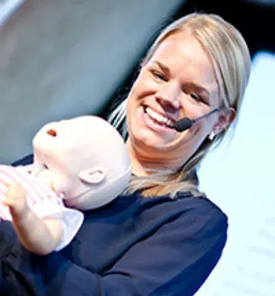 Camilla Thurell föreläser på scen med ett stort leende på läpparna och en bebisdocka i händerna.