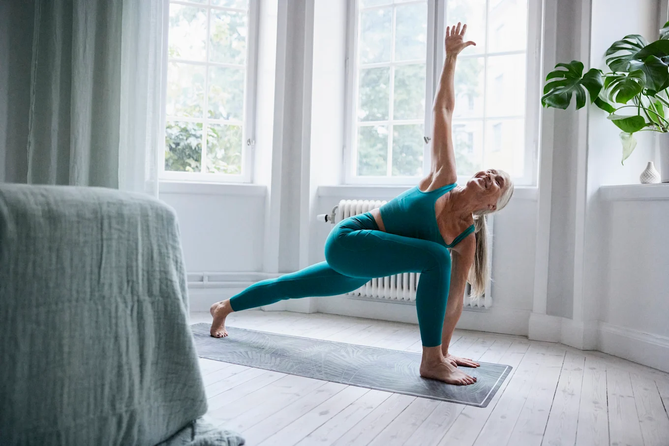 Kvinna står i yogapose med en arm i luften och den andra bredvid sin vänstra fot. Den högra foten är stäckt bakåt. Hon är i ett lugnt och luftigt sovrum med utsikt mot grönska. 