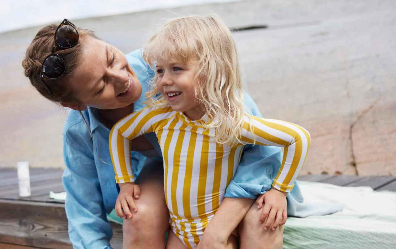 En glad kvinna sitter ner på en bänk vis stranden och kramar om ett barn i baddräkt.
