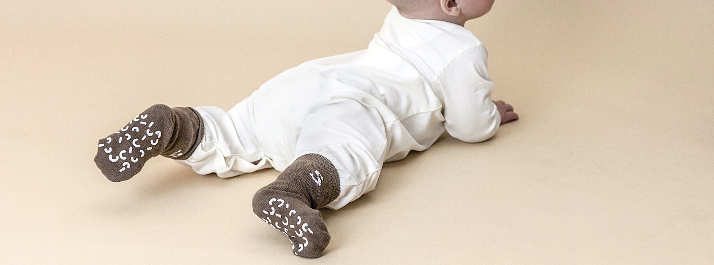 Bebis i vit sparkdräkt ligger på mage på beige underlag och bär bruna halksockor med vita mönster.