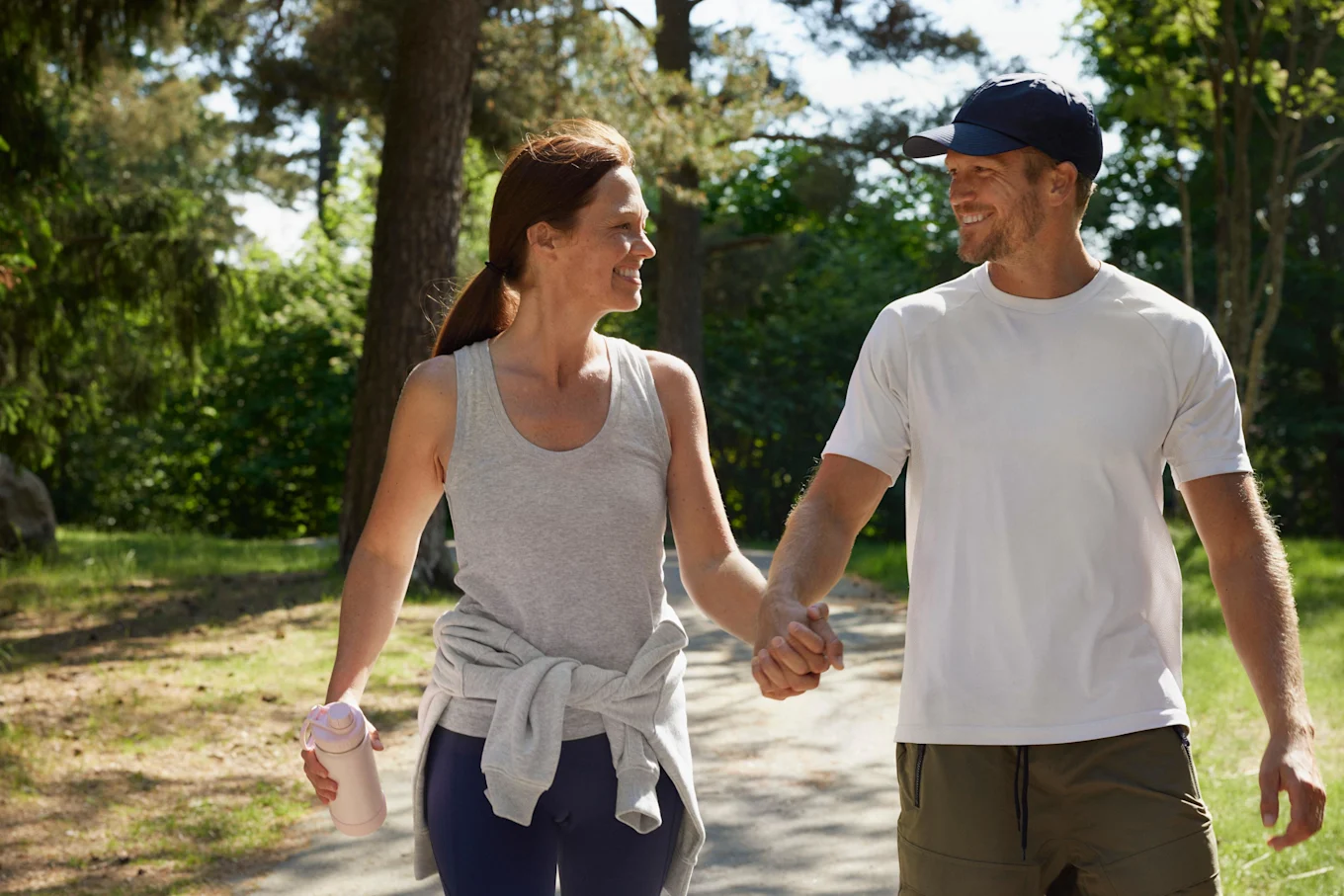 En kvinna och man är ute och går på en promenad i en solig, öppen skog. De håller varandra i handen och tittar på varandra och ler. Kvinnan håller i en vattenflaska och det ser ut som att det är varmt ute. 