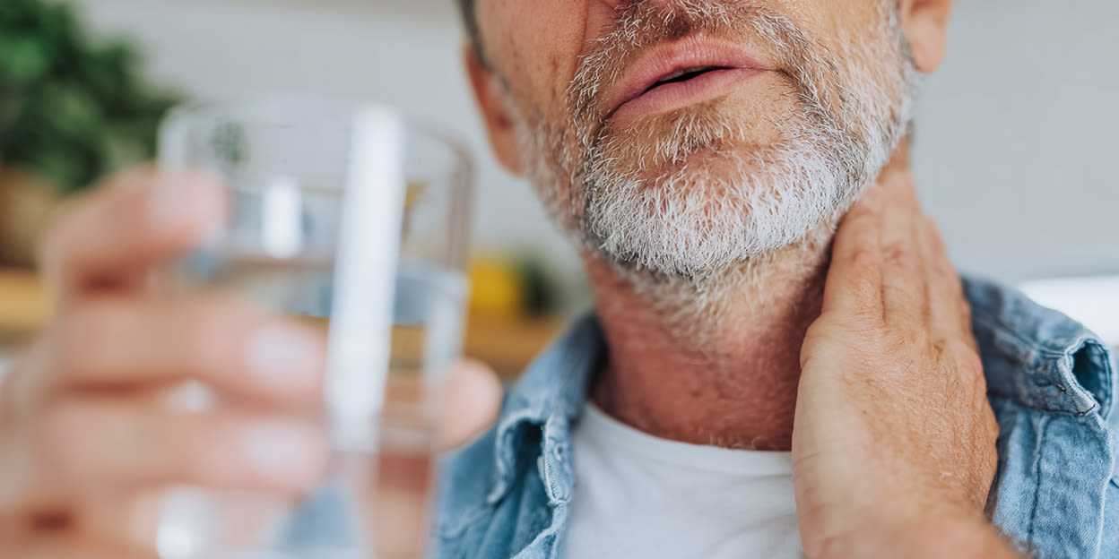 Man håller ett glas vatten i ena handen och den andra på sin onda hals.