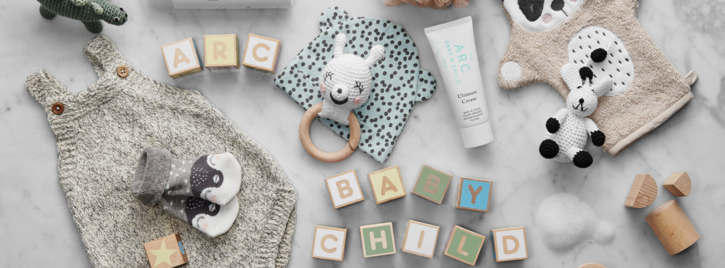 Stickad barnklänning, babysockor, träklossar med texten "ARC BABY CHILD", babykräm, gosedjur och leksaker på marmorbord.