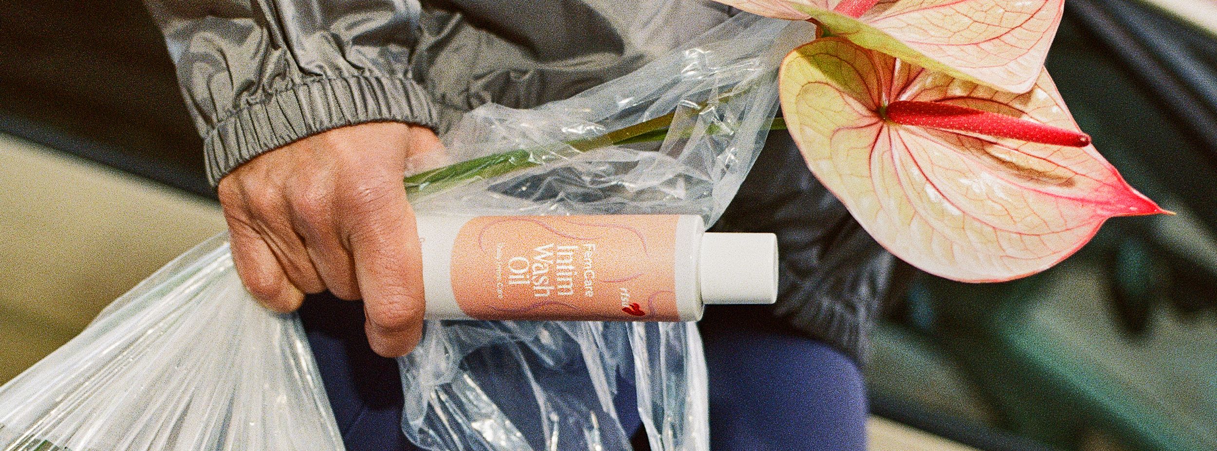 En hand håller en plastpåse med en rosa blomma och en flaska märkt "FemiCare Intim Wash Oil" mot suddig bakgrund.