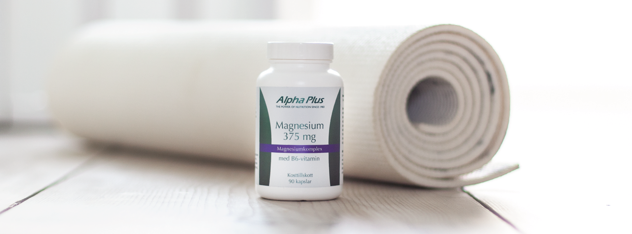 Burk med Alpha Plus Magnesium 375 mg kosttillskott står på golvet bredvid en hoprullad yogamatta i ljust rum.