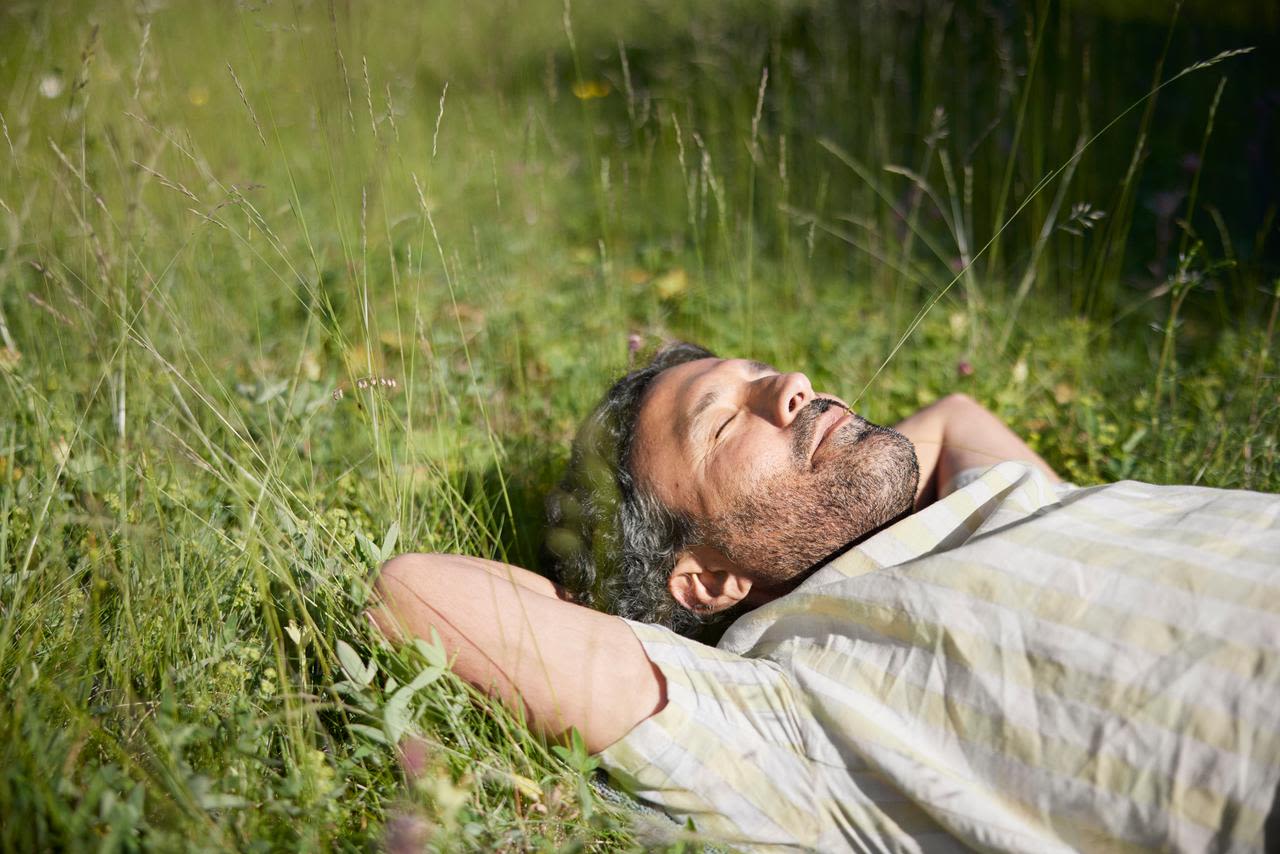 Man i gul och vitrandig skjorta ligger med stängda ögon på en sommaräng med ett grässtrå i mungipan och njuter av solskenet. 