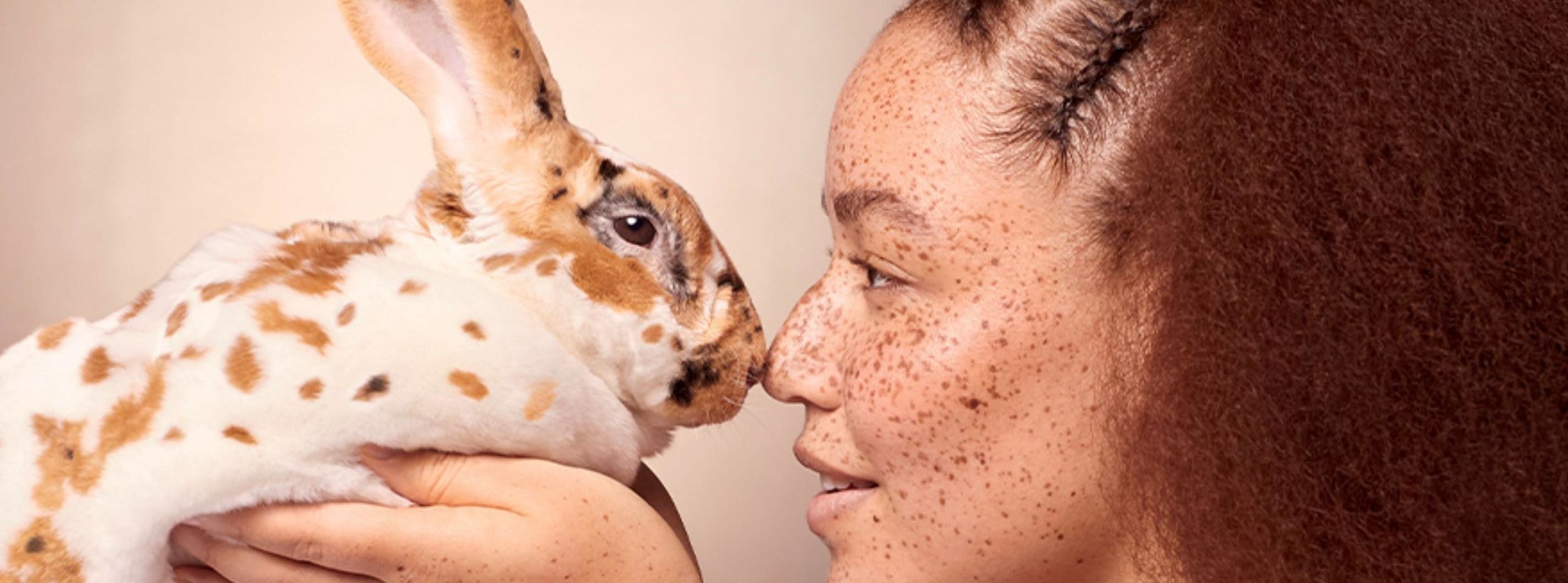 Kvinna med fräknar håller en vit och brunfläckig kanin, deras näsor nästan nuddar varandra mot en ljus bakgrund.