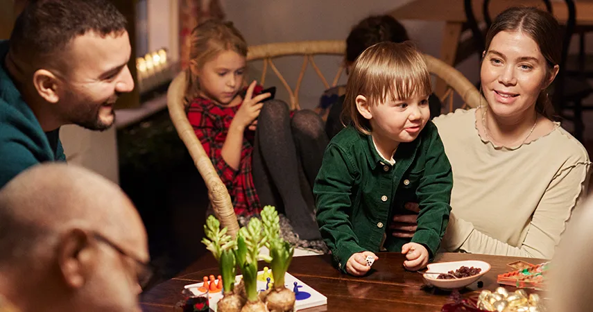 En familj med barn och vuxna sitter samlade runt matbordet och pratar, rummet är pyntat för jul med hyacinter och ljusstakar. På bordet står julgodis.