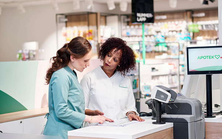 Två kvinnliga apotekare står vid kassan på Apotek Hjärtat och diskuterar.
