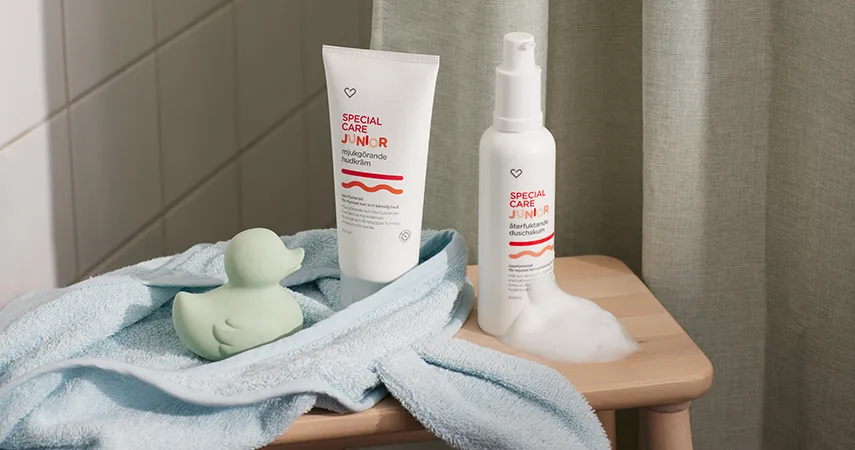 På en pall i badrummet ligger en barnmorgonrock och en grön gummianka. Bredvid står två av Apotek Hjärtats produkter inom Special Care – Junior.